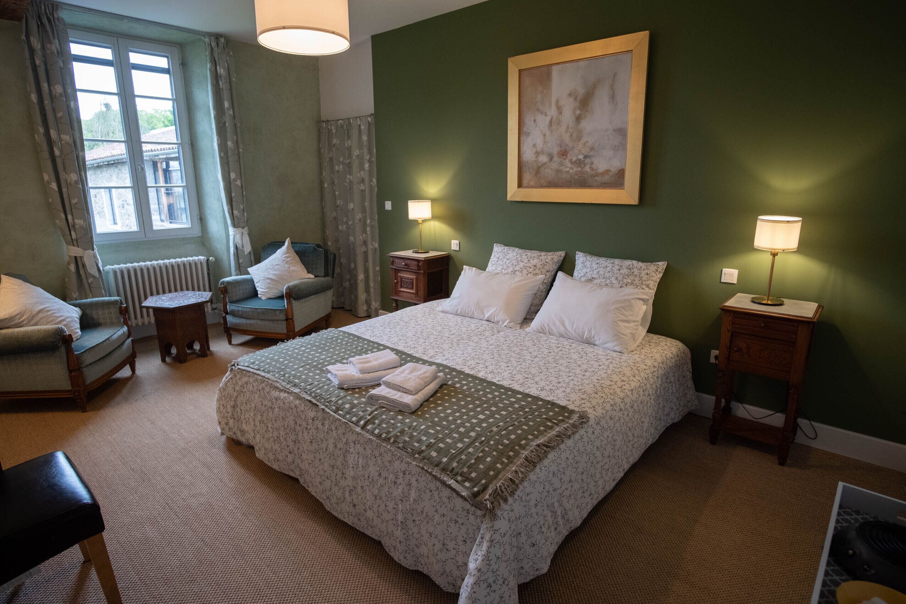 Habitación de hotel con una cama grande, dos mesitas de noche con lámparas, una silla, una pequeña mesa y un radiador.