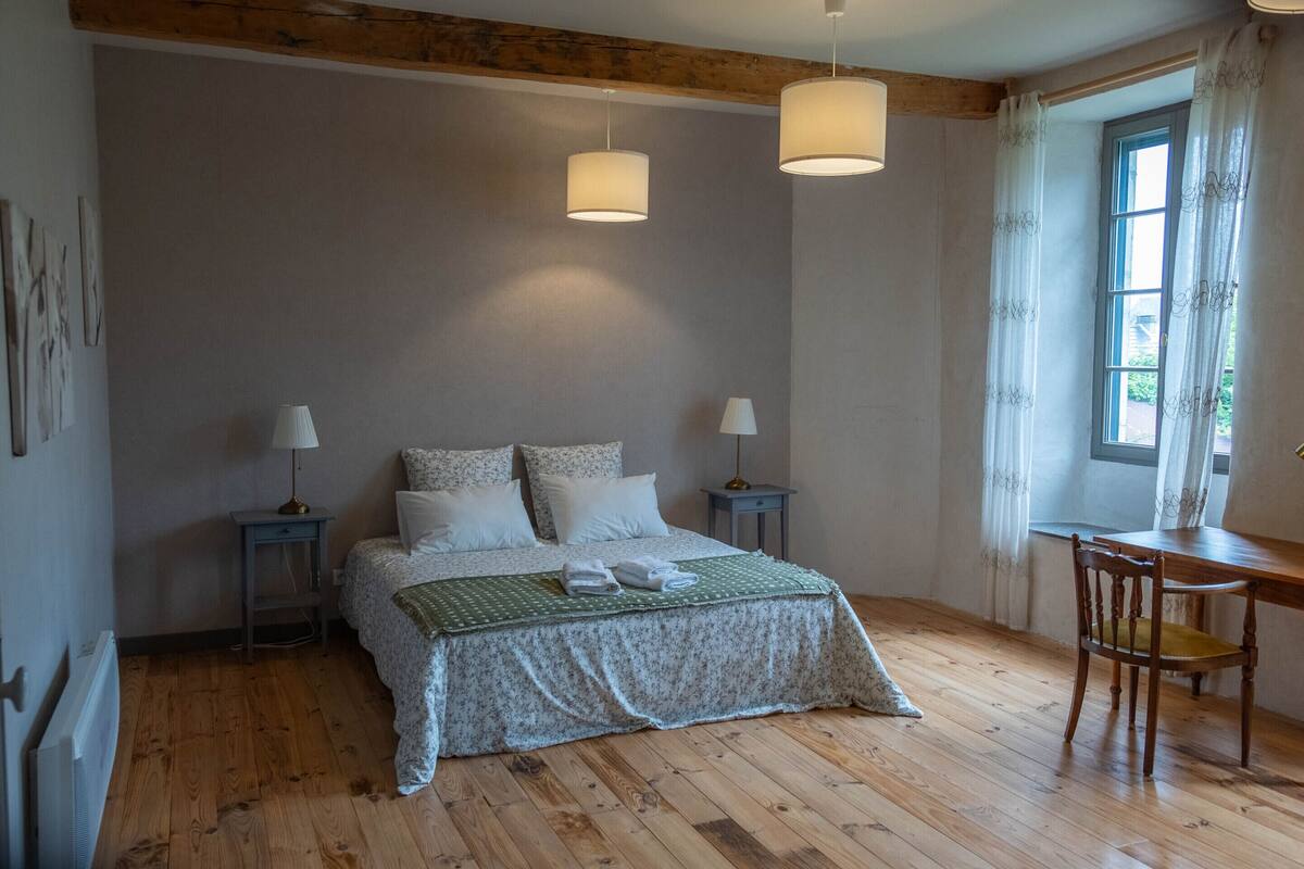 Un dormitori amb sòl de fusta, un llit amb col·cha de flors verd i blanc, dues làmpades de nit, un escriptori de fusta amb una cadira, i una finestra amb cortines.