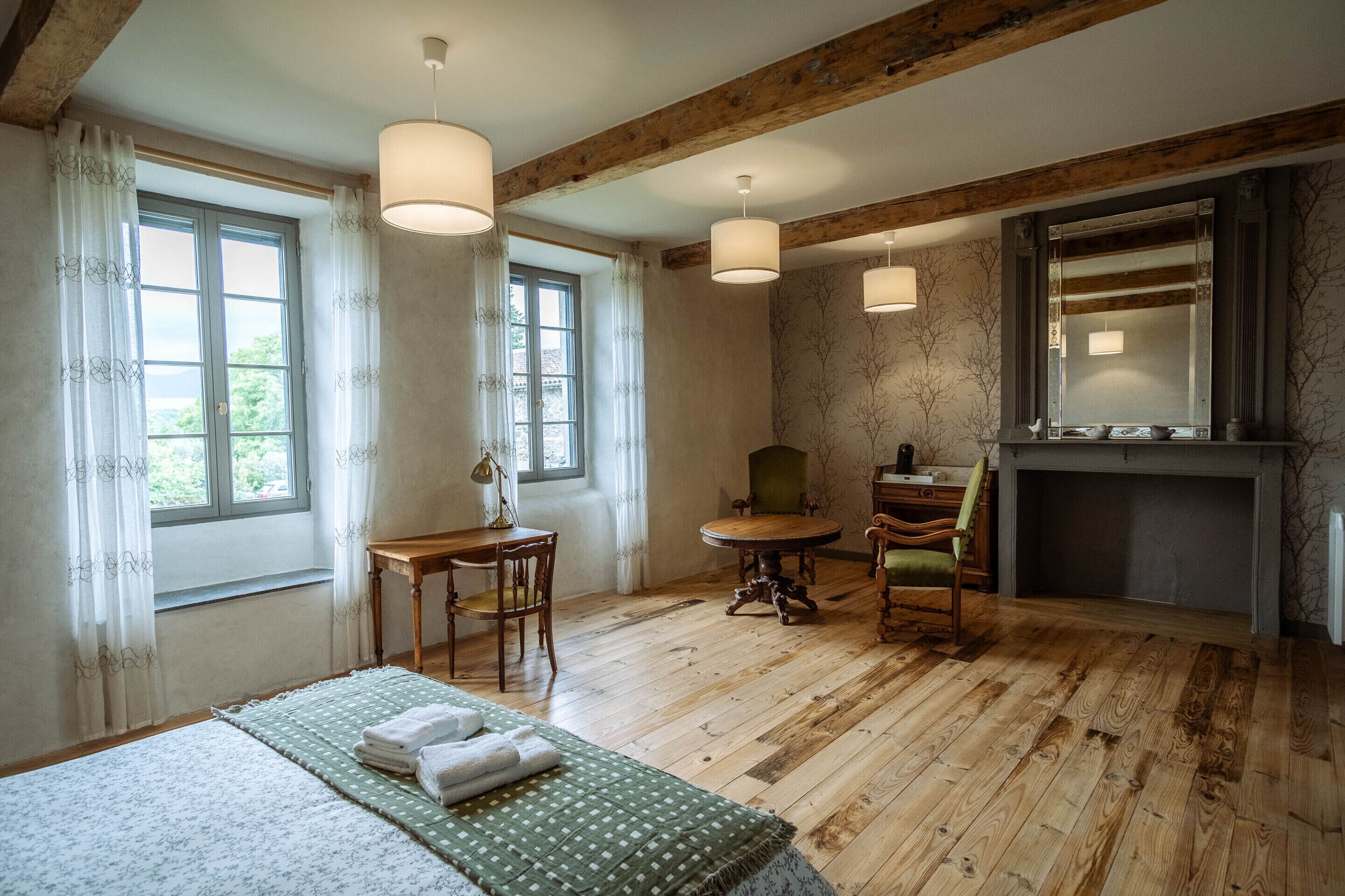 A room with wooden floors, a bed with a green and white patterned bedspread, a wooden table with a chair, a fireplace, and two hanging lamps.