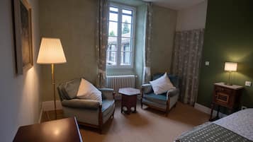 Una habitación con dos sillones, una mesita, una cama, un radiador y una ventana con cortinas.