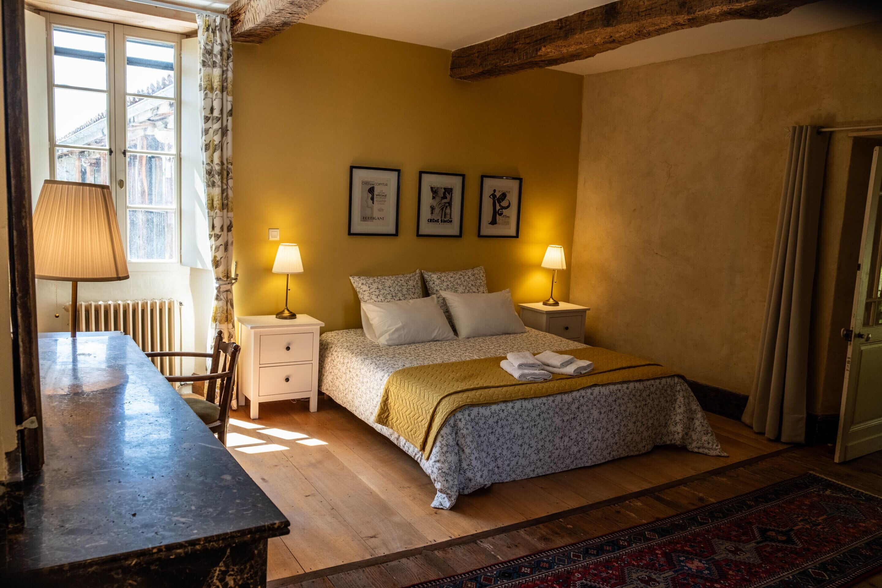 A bedroom with a bed, two bedside tables, a lamp, a chair, a radiator, and a window with curtains.