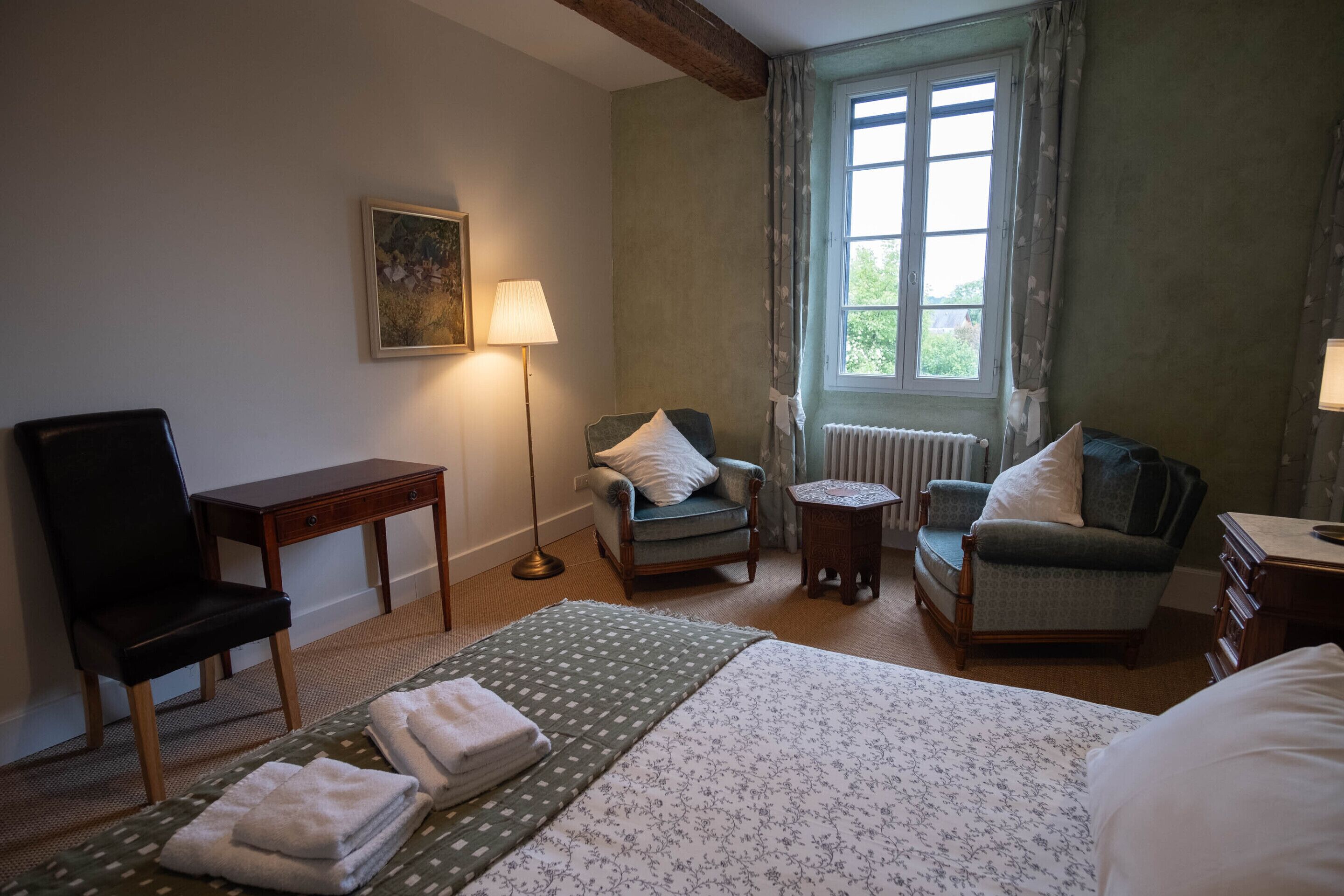 Een hotelkamer met een bed, twee fauteuils, een klein tafeltje, een lamp, een schilderij aan de muur en een raam met gordijnen.