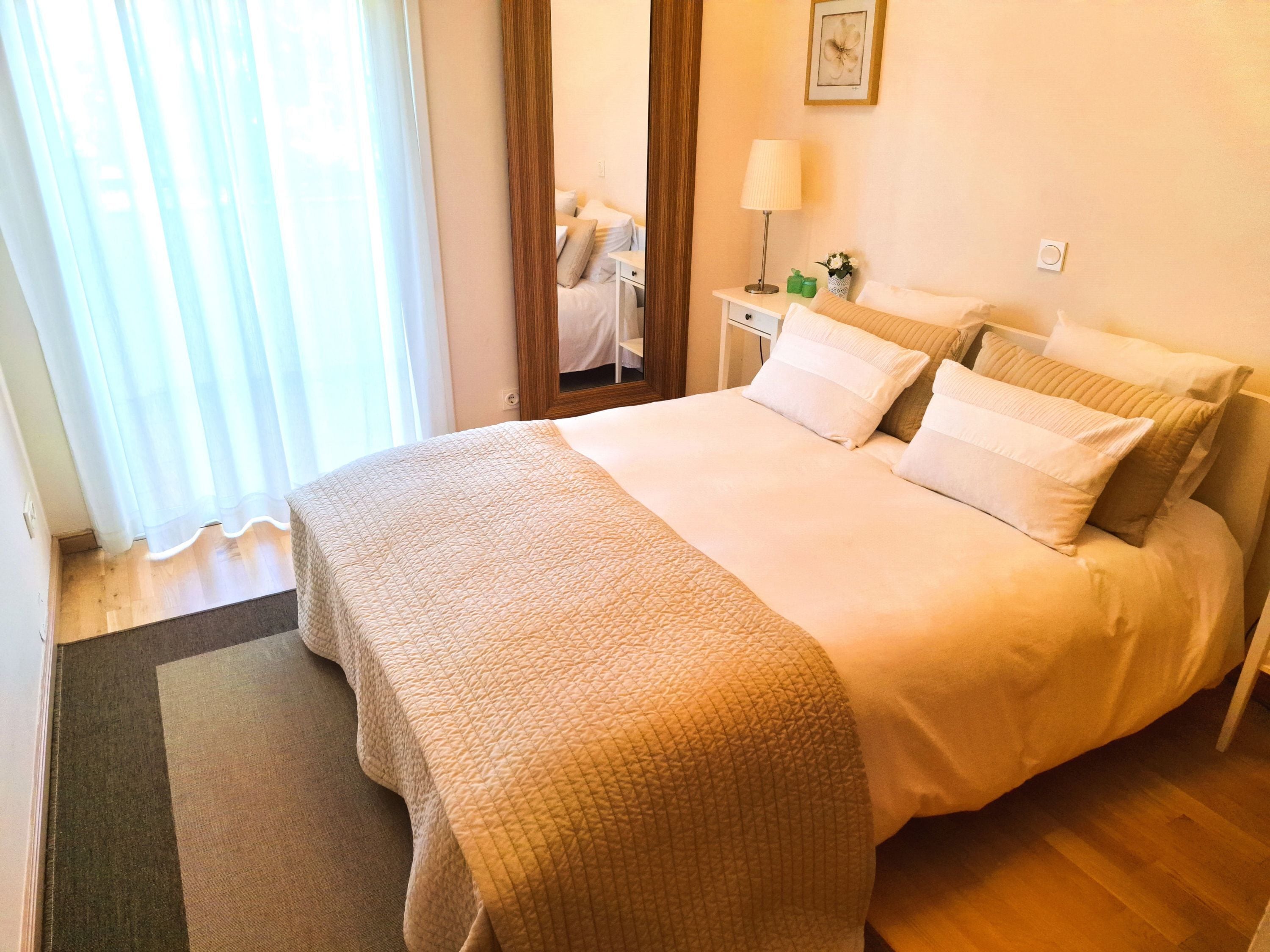 A neatly made bed with a quilted bedspread and pillows, a bedside table with a lamp and a small plant, and a window with sheer curtains.