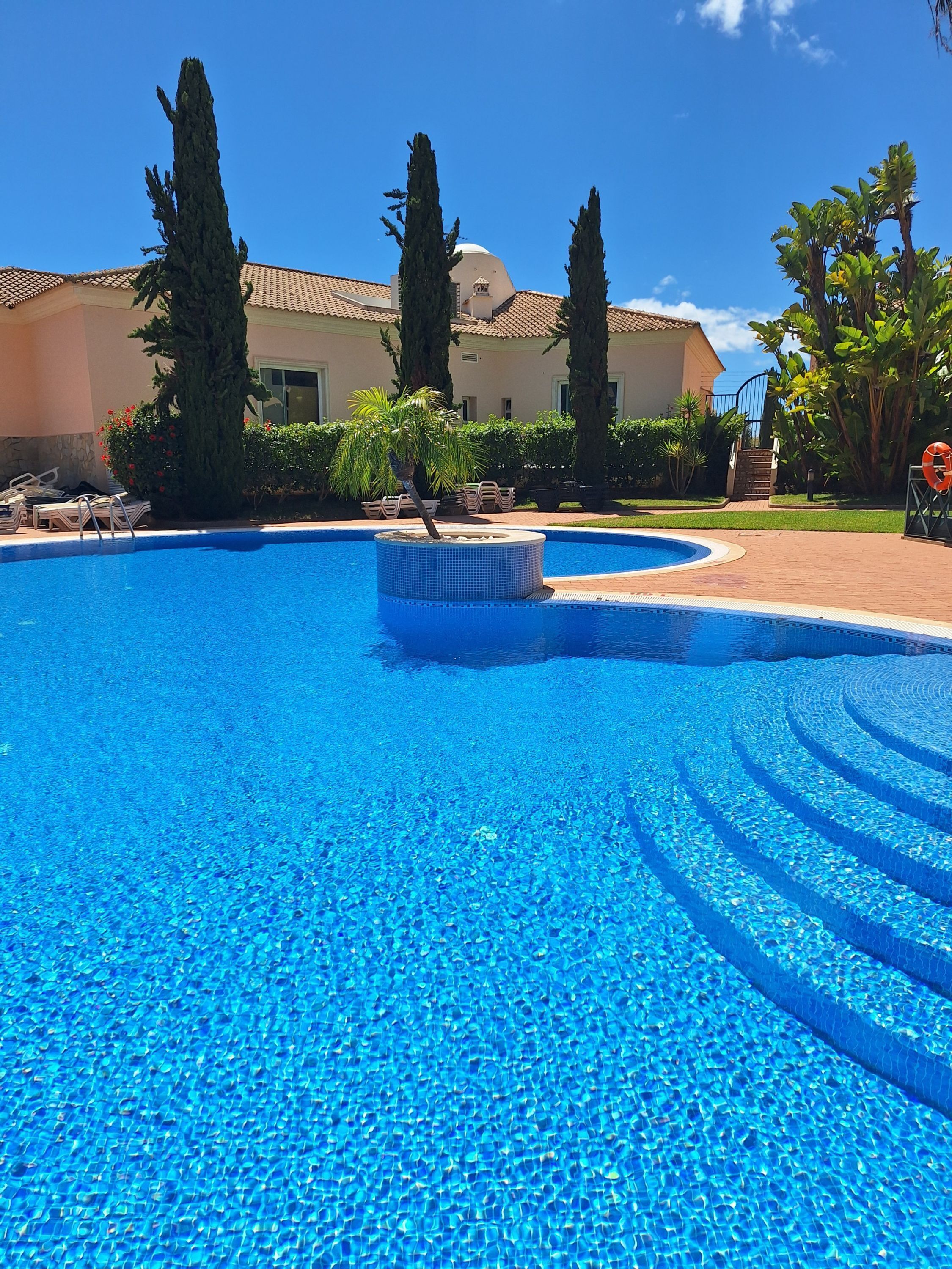 Una piscina azul cristalina con un bordeado de baldosas, rodeada de palmeras y un edificio con techo de tejas.