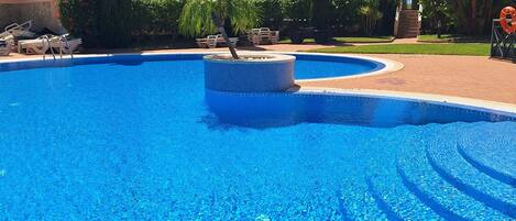 Una piscina azul cristalina con un bordeado de baldosas, rodeada de palmeras y un edificio con techo de tejas.
