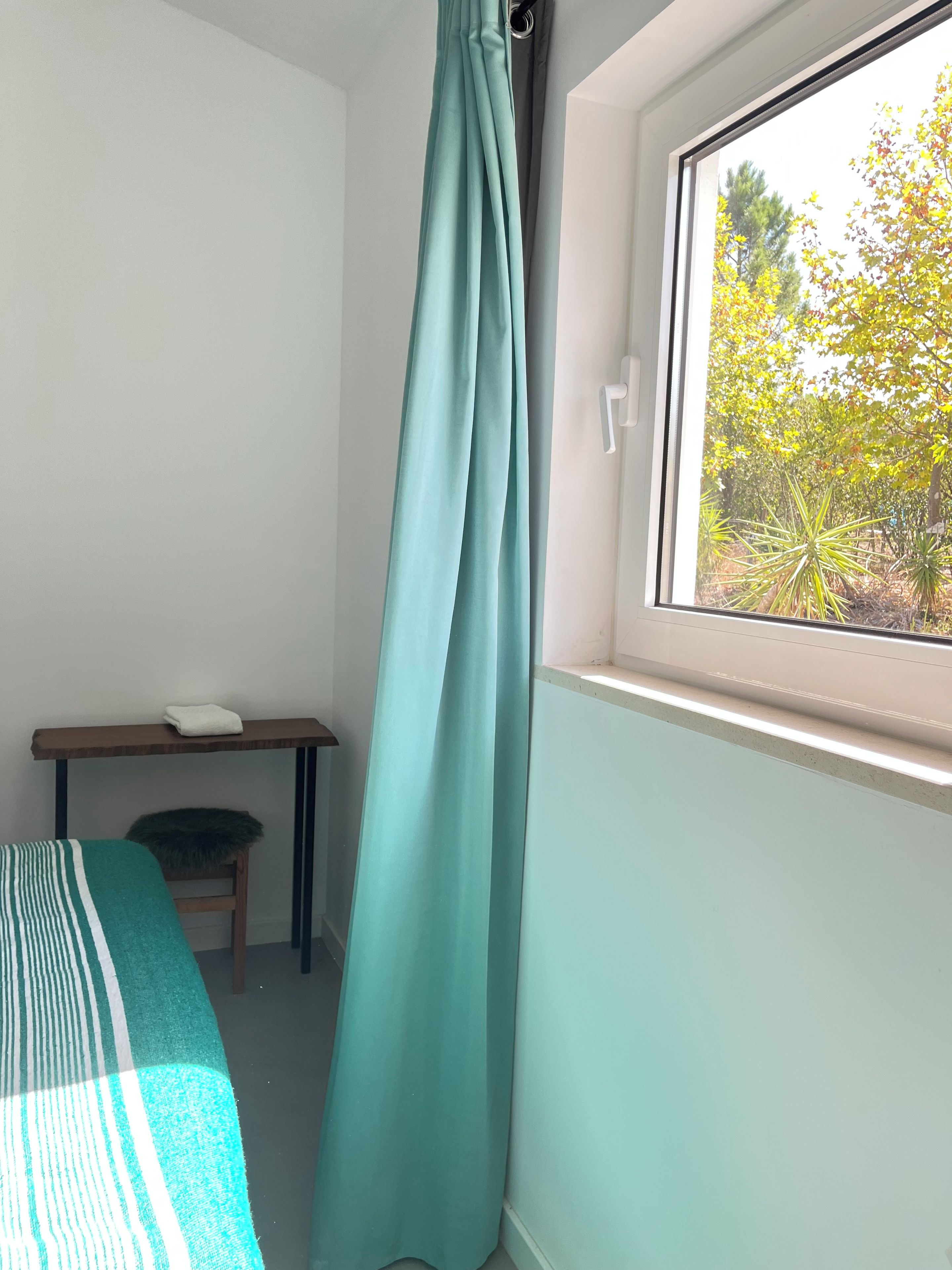 A room with a window, a bed with a striped blanket, a small table with a white object, and a curtain.