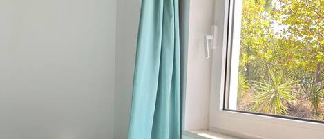 A room with a window, a bed with a striped blanket, a small table with a white object, and a curtain.