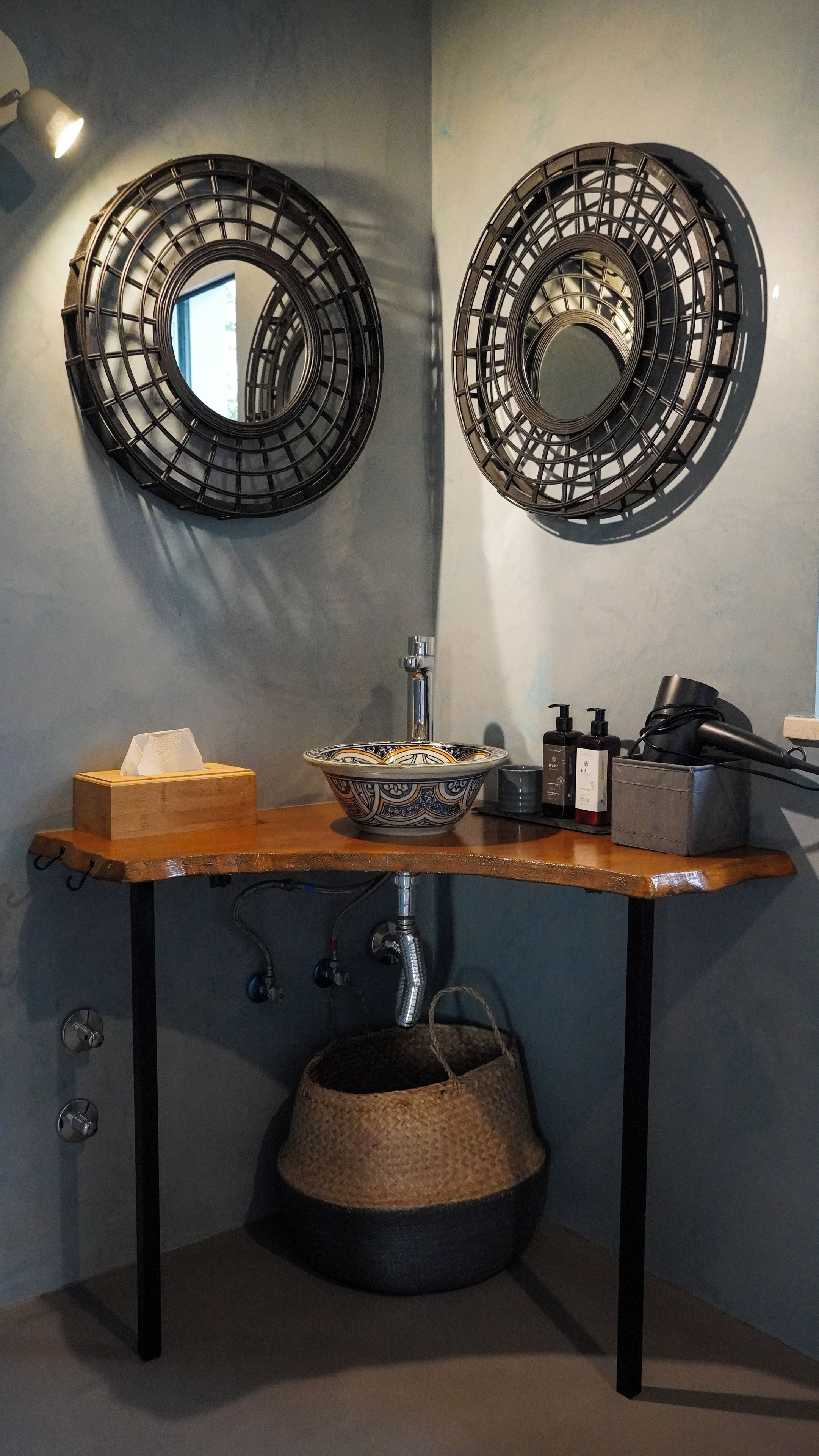 A bathroom with a wooden vanity, a sink with a decorative bowl, two wall-mounted mirrors, a hanging basket, and a towel dispenser.