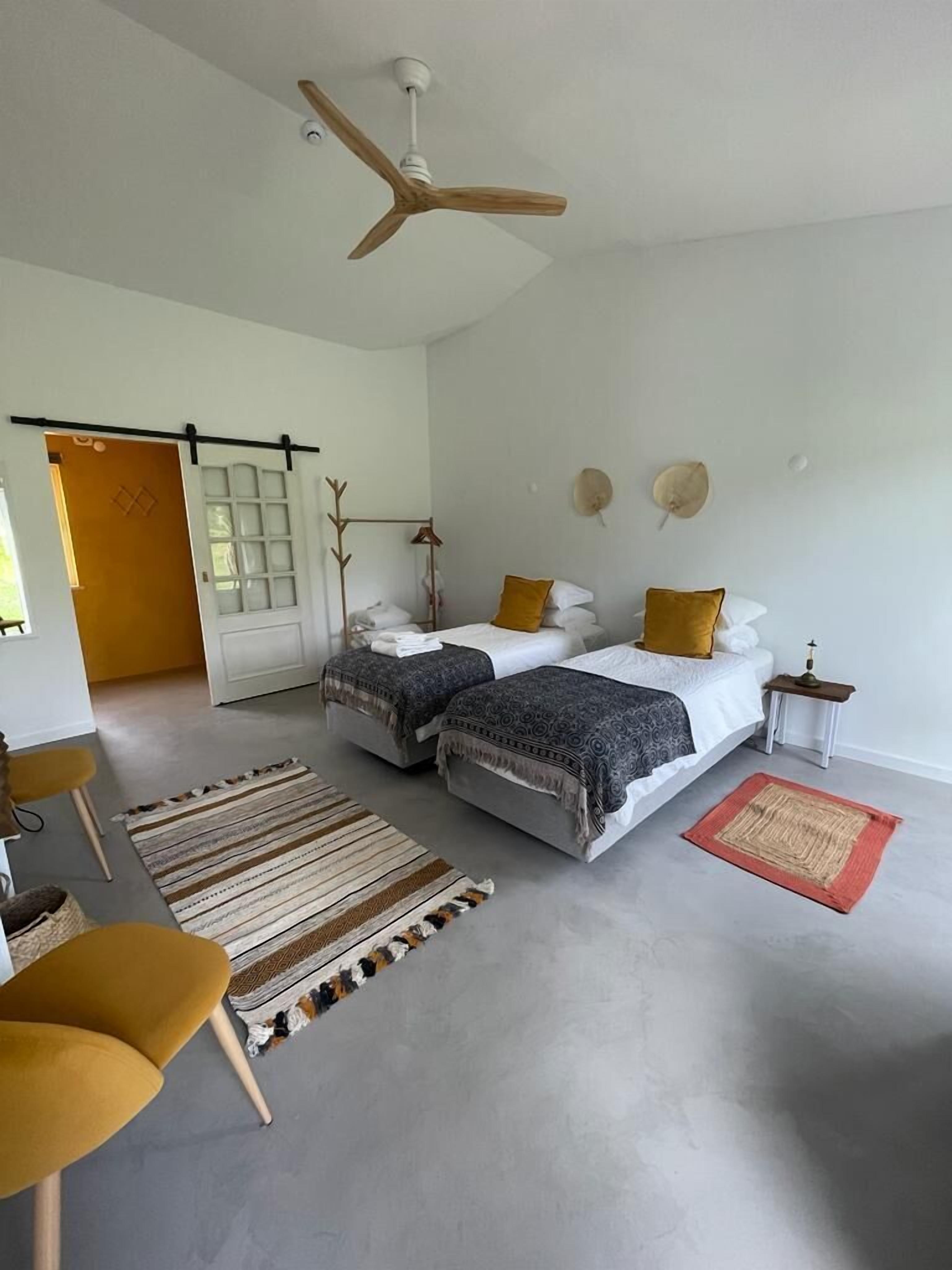 Une chambre moderne avec deux lits, un ventilateur de plafond, une porte en bois et une petite table avec une lampe.