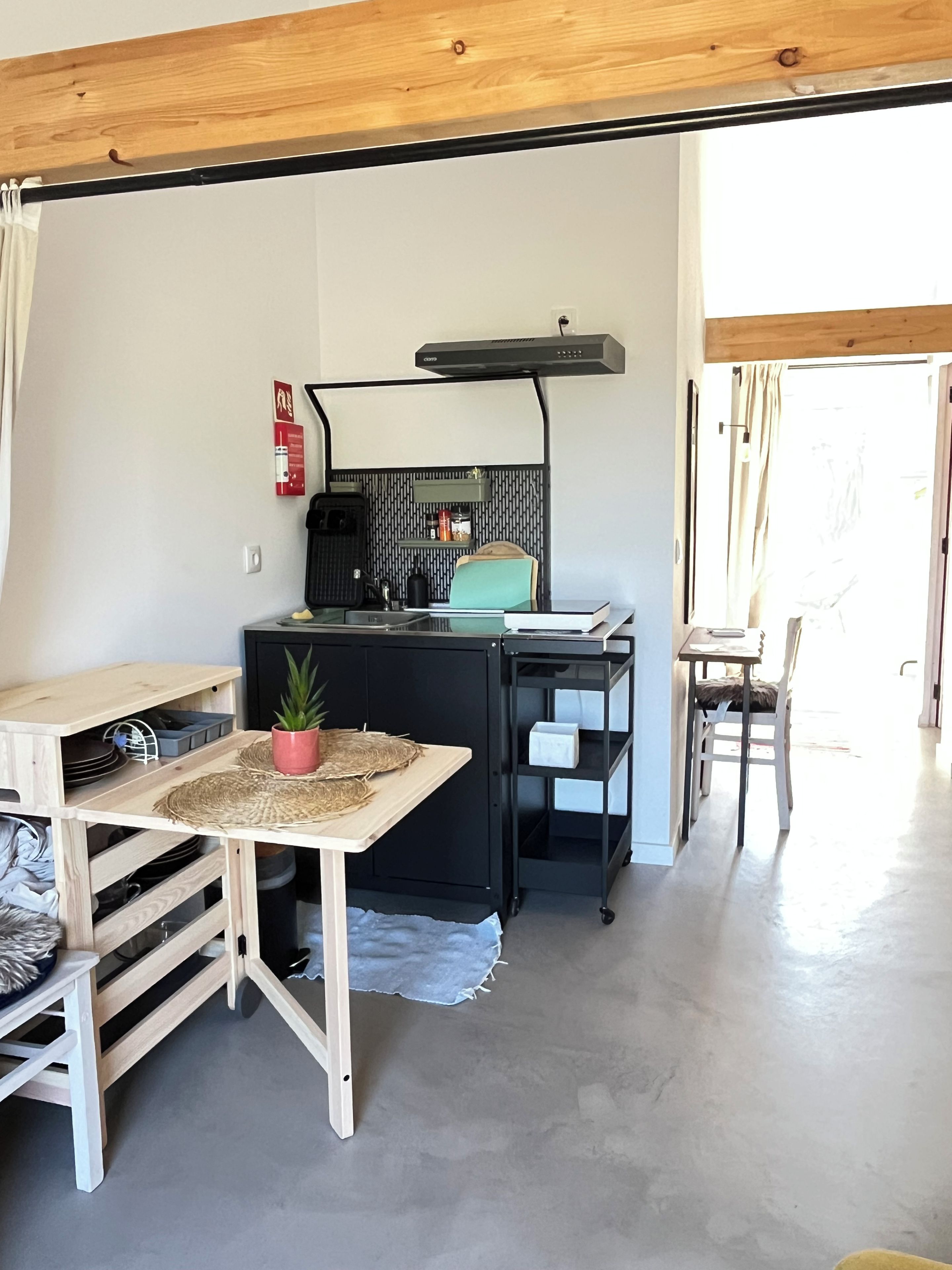 Een compacte keuken met een zwart aanrecht, een koffieapparaat en een klein eiland met een stoel. Er hangt een brandblusser aan de muur en er staat een plant in een pot op het aanrecht.