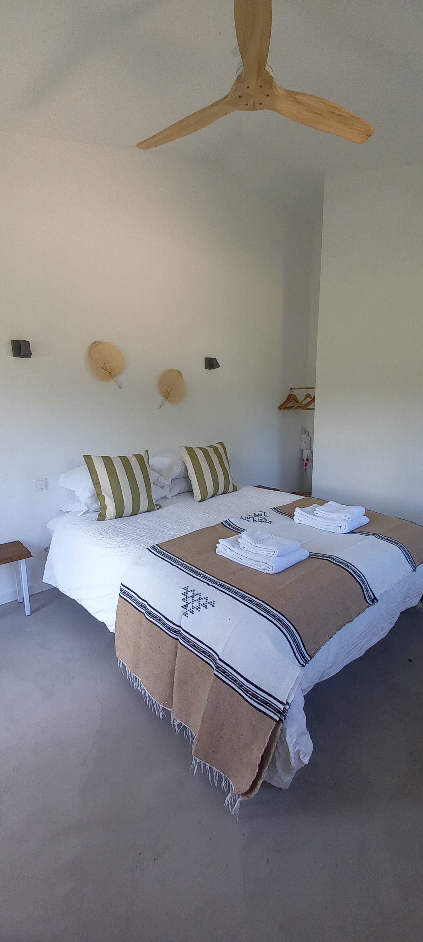 Un dormitorio con cama, almohadas, manta, mesita de noche y ventilador de techo.