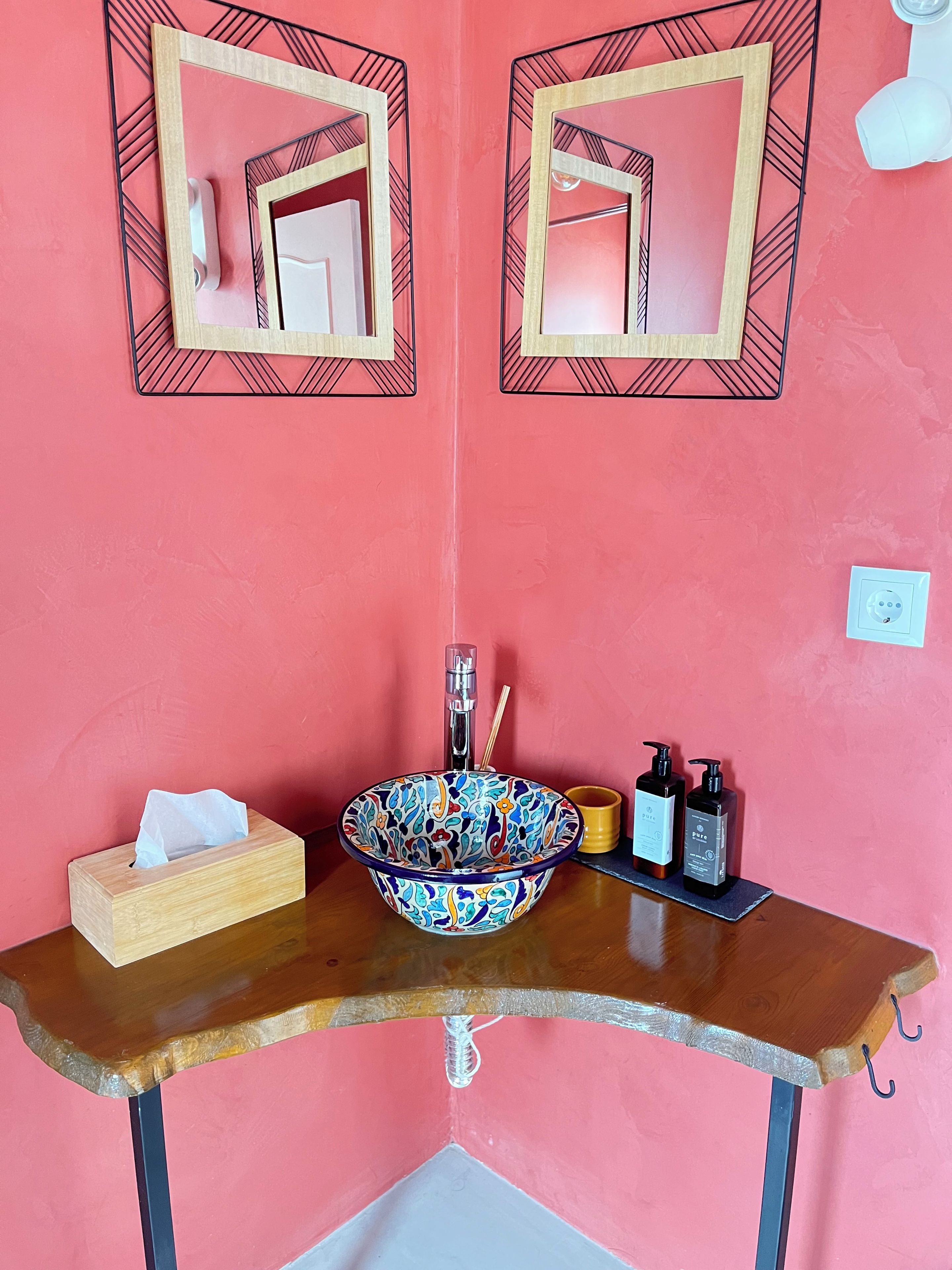 Une salle de bain avec un mur rose, deux miroirs, un lavabo avec un récipient coloré, une boîte à mouchoirs, un distributeur de savon et un porte-brosse à dents.
