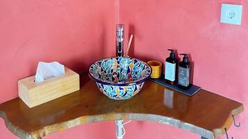 Une salle de bain avec un mur rose, deux miroirs, un lavabo avec un récipient coloré, une boîte à mouchoirs, un distributeur de savon et un porte-brosse à dents.
