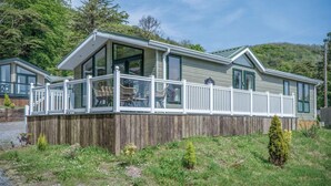 Ein eingeschossiges Mobilheim mit überdachter Veranda, großen Fenstern und einer Holzterrasse.
