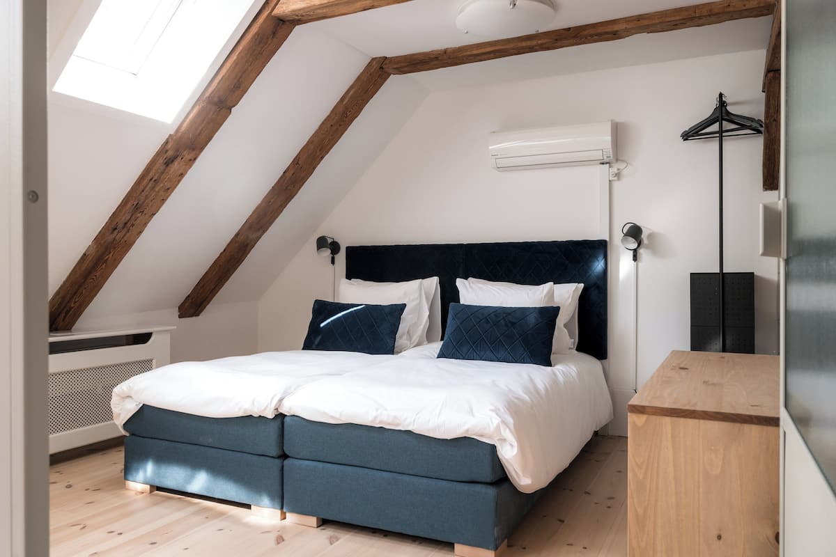 Ein Schlafzimmer mit schrägen Wänden, einem Bett mit dunklem Kopfteil, einem Nachttisch und einer Pendelleuchte.
