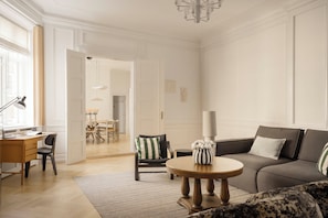Ein Wohnzimmer mit einer Couch, einem runden Couchtisch, einem Schreibtisch und einem Sessel. Eine Tür führt in einen anderen Raum.