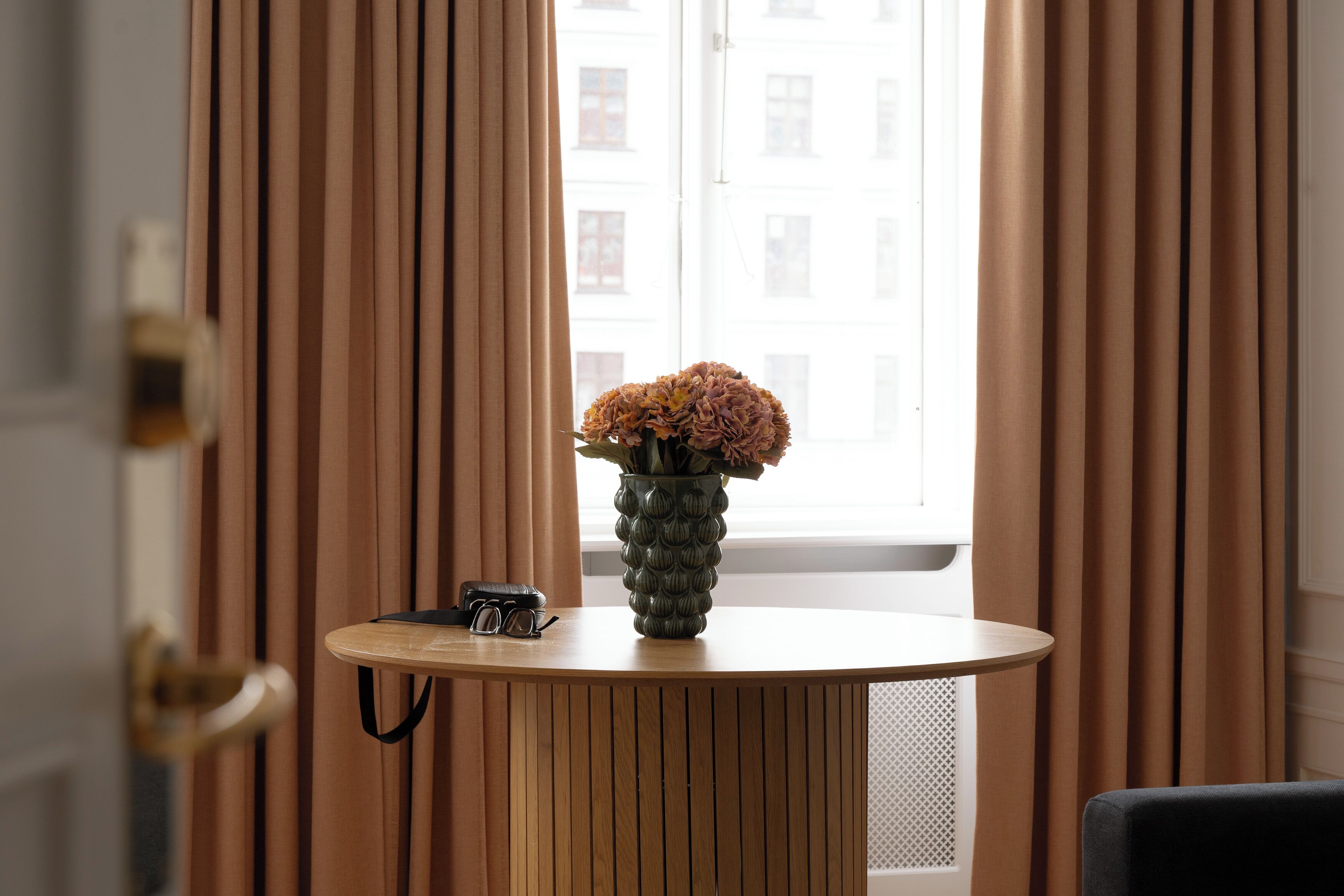 Ein Zimmer mit einem Holztisch, einem Blumenstrauß in einer Vase und einem Vorhangfenster.