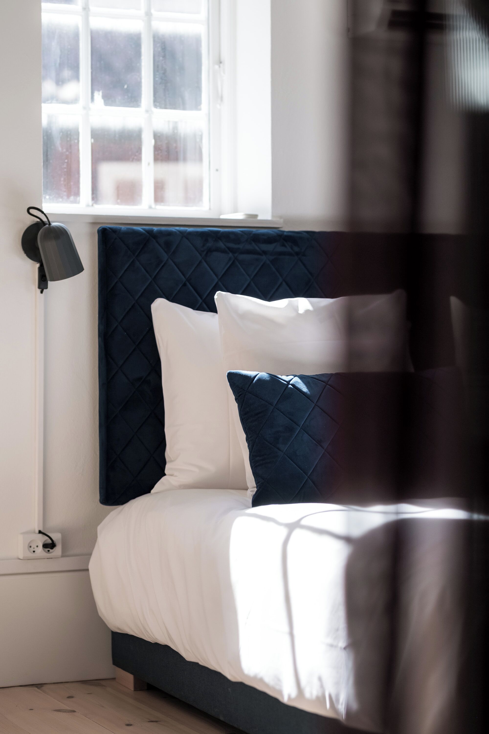 A neatly made bed with a quilted headboard, a bedside lamp, and a window with a view of the outside.