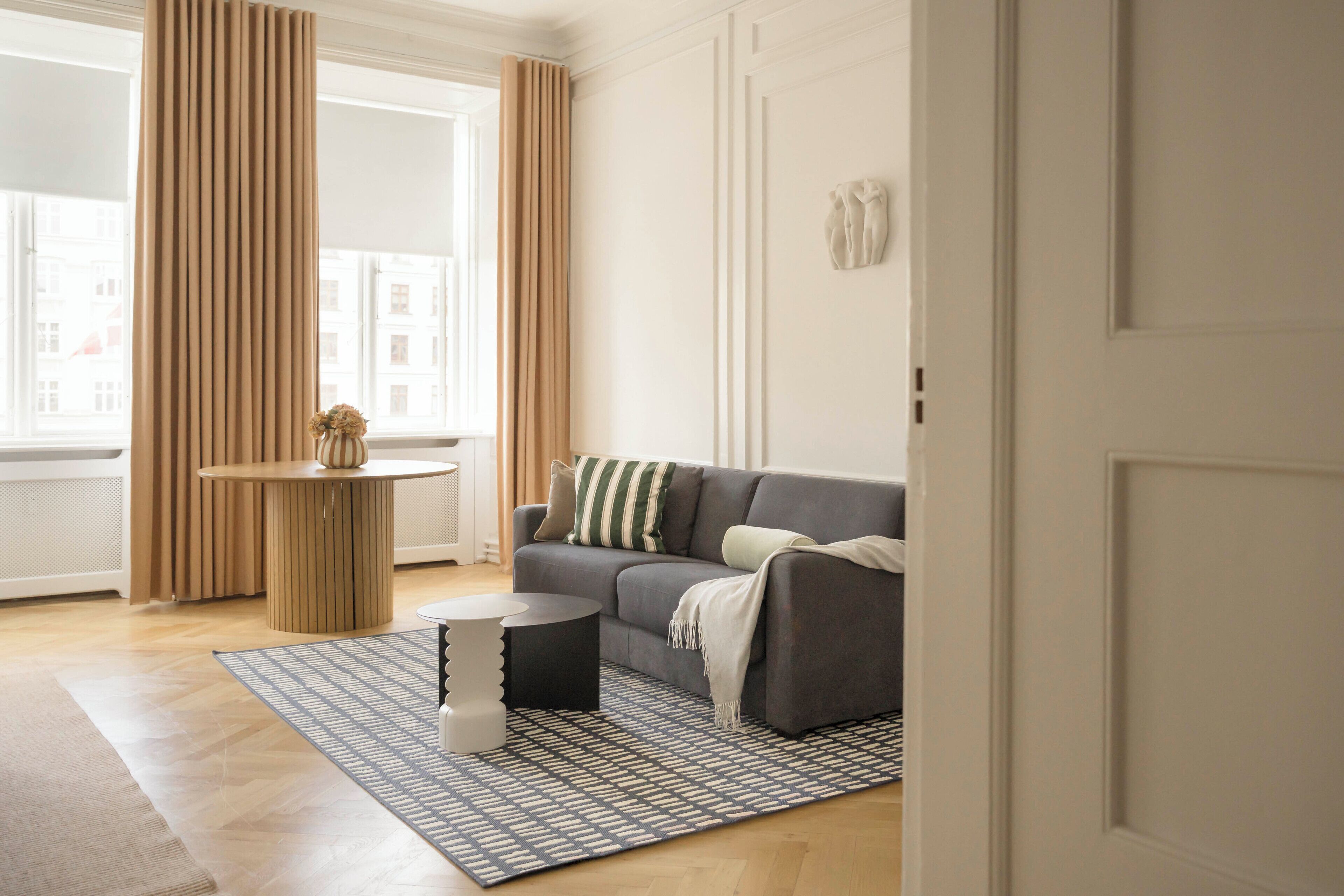 Una sala de estar moderna con un sofá gris, una mesa redonda de centro, una alfombra estampada y una ventana con cortinas.
