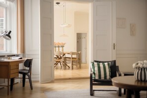 Ein Zimmer mit Schreibtisch, Stuhl, einem Esstisch mit Stühlen, einer Vase mit Blumen und einer Tür zu einem weiteren Raum.