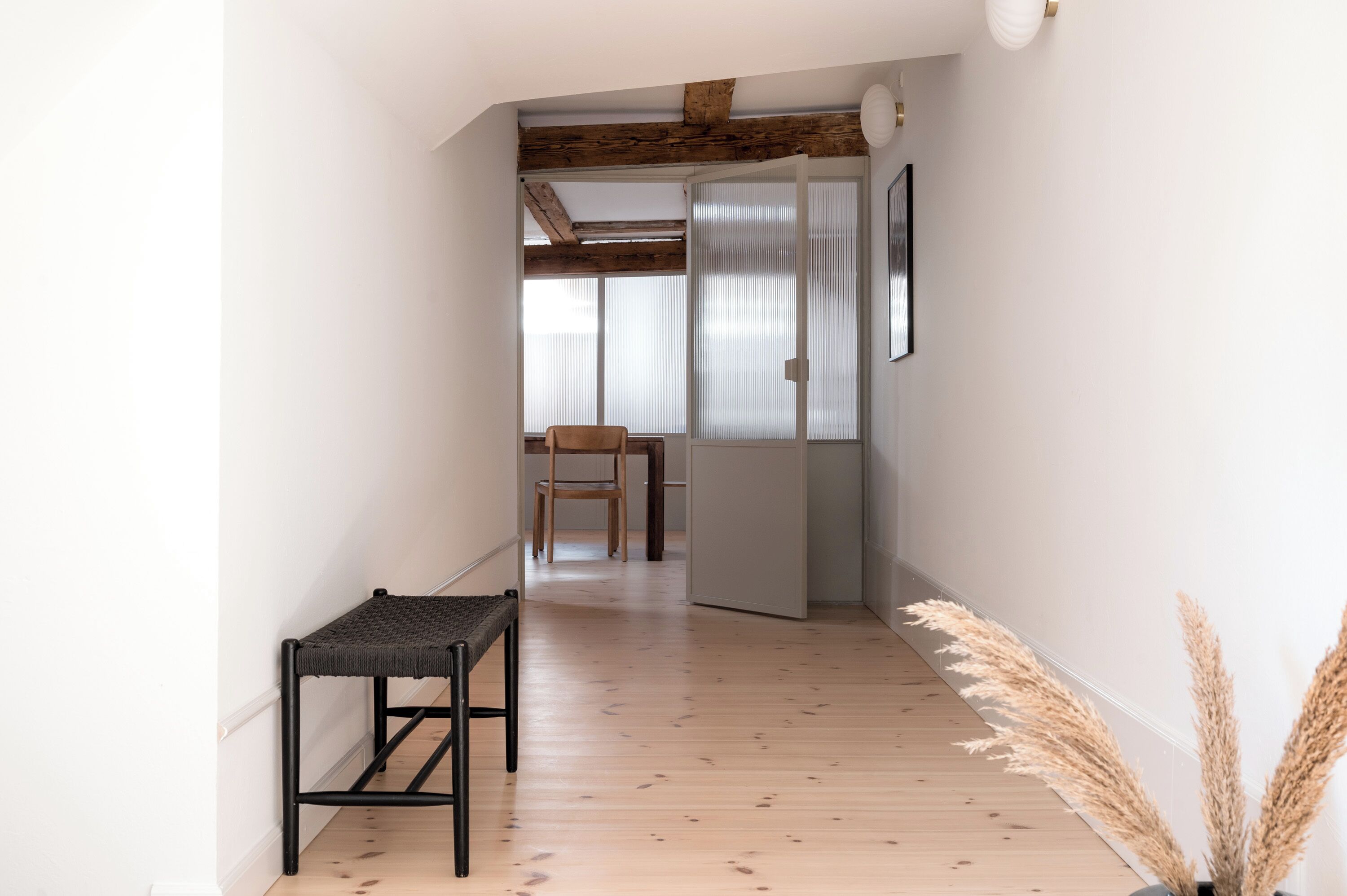 Un pasillo con suelo de madera, un banco, una silla y una puerta que da a otra habitación.