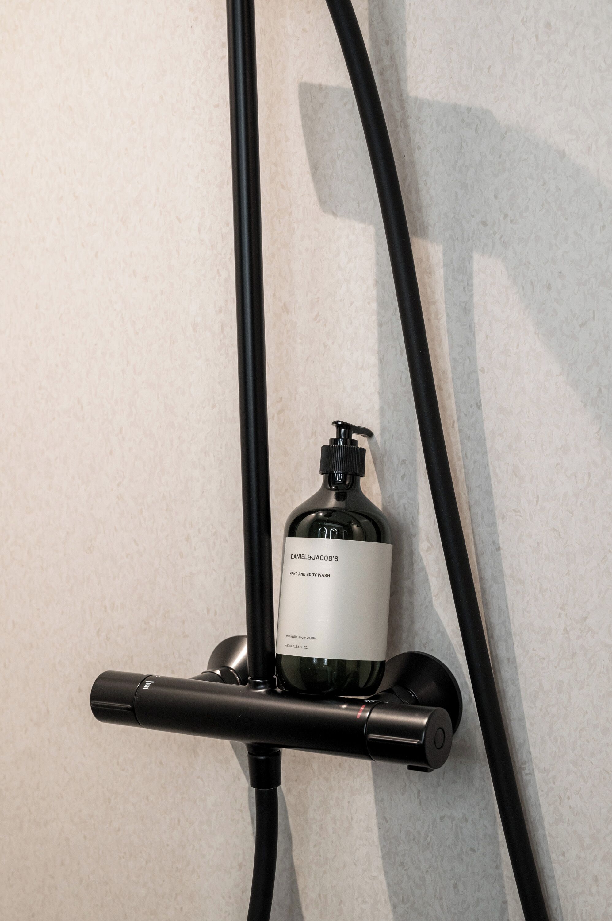 A black bottle of Daniel Jacob's body wash mounted on a wall with a black hose.