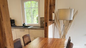 Een houten eettafel met stoelen, een raam met uitzicht op groen, en een lamp met gedroogde planten.