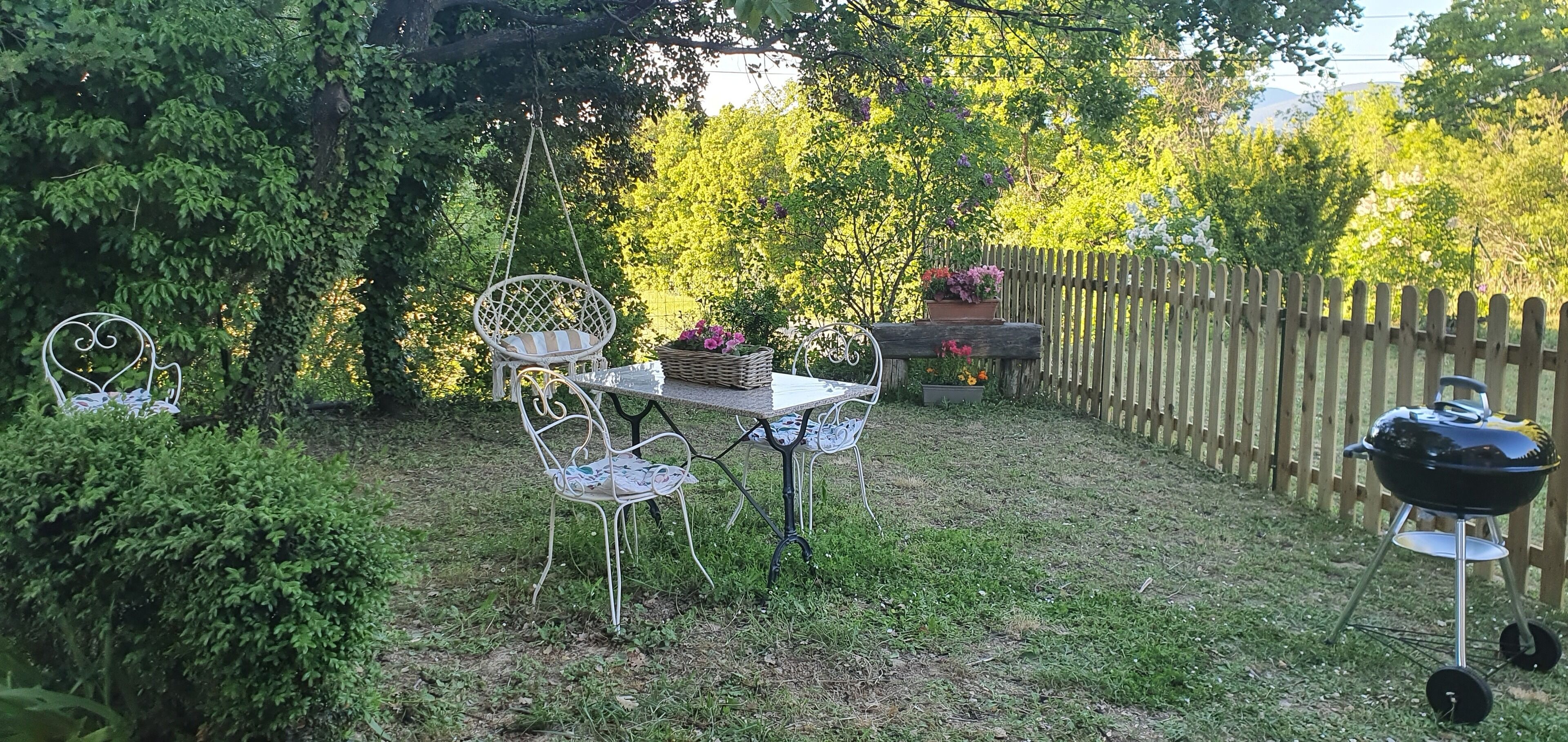 Un jardin avec une clôture en bois, une table avec des chaises, un fauteuil suspendu et un barbecue.
