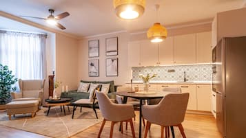 Una sala de estar moderna con un sofá, sillas, una mesa de centro y una mesa de comedor. Se observa una zona de cocina con armarios y un refrigerador.