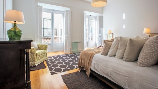A bedroom with a bed, two bedside lamps, a green table lamp, a patterned rug, and a view of the outside balcony. - Portas de São Bento Apartment (Lisbon)