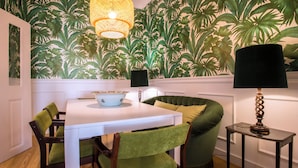 A dining area with a white table, green upholstered chairs, a green sofa, a wall with a tropical leaf pattern, and a hanging light fixture. - Portas de São Bento Apartment (Lisbon)