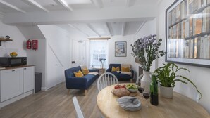 Una cocina moderna con microondas, una mesa de comedor dispuesta con platos, cubiertos y una botella de vino. En la zona de estar, hay un sofá azul con cojines amarillos, y una ventana con vistas a la ciudad.