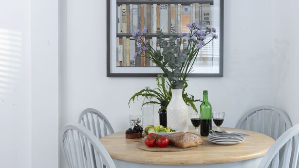 Una mesa redonda para comer con una silla blanca, un florero con flores, una botella de vino y una hogaza de pan.