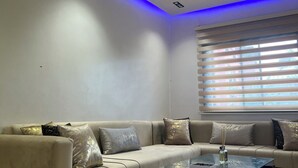 Un salon moderne avec un canapé d’angle beige, une table basse en verre et un plafond doté d’un éclairage LED bleu.