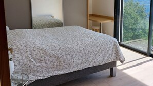 Une chambre avec un grand lit, un plancher en bois, un bureau, une chaise, un miroir et une vue sur les montagnes par une grande fenêtre.