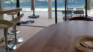 Un espace repas moderne avec une grande table en bois, des tabourets de bar et une vue sur les montagnes et un lac, grâce à de grandes fenêtres.