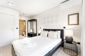 Quarto de hotel moderno com uma cama grande, uma mesinha de cabeceira com abajur, um espelho fixado na parede e um quadro emoldurado.