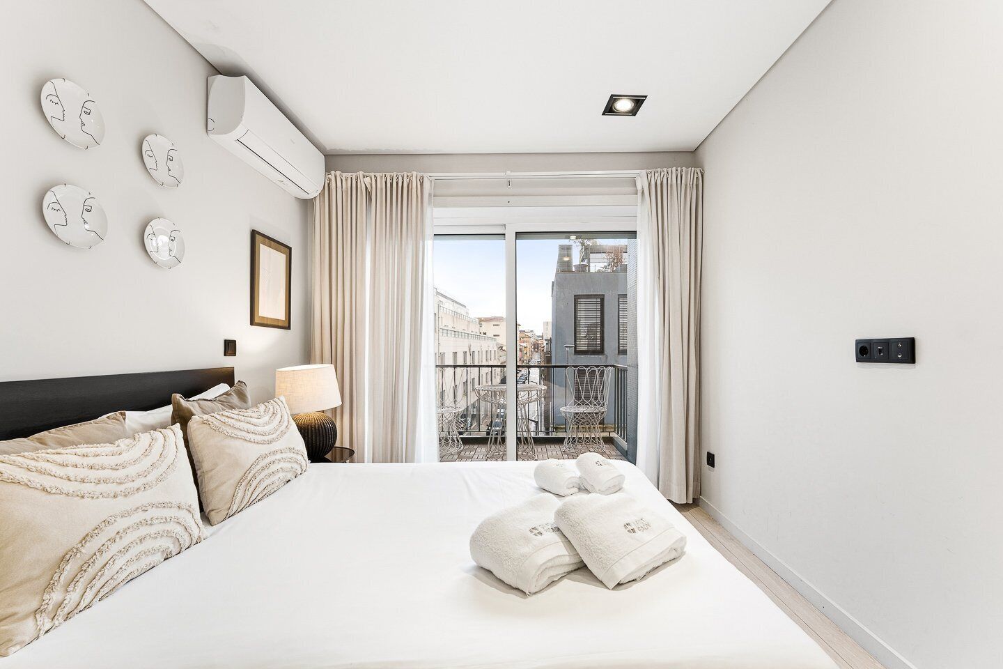 A hotel room with a large bed, a bedside table with a lamp, a view of the city through a balcony door, and a wall-mounted air conditioning unit.