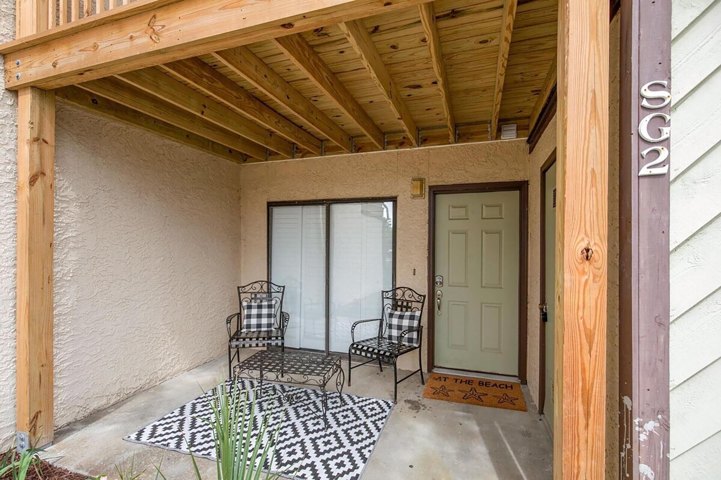 A covered patio with two metal chairs and a patterned rug, a wooden door, and a mailbox.