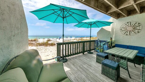 Une terrasse en bois avec une table et des chaises, deux grands parasols et une vue sur la plage.