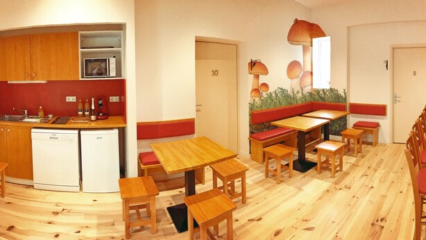 A compact kitchen with wooden cabinets and a microwave, a dining area with wooden tables and red cushioned benches, and a wall with a mushroom mural. - Défiplanet' (Dienne)