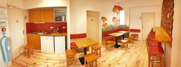 A compact kitchen with wooden cabinets and a microwave, a dining area with wooden tables and red cushioned benches, and a wall with a mushroom mural. - Défiplanet' (Dienne)