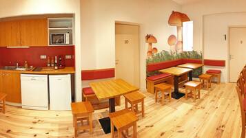 Una cucina compatta con mobili in legno e un forno a microonde, una zona pranzo con tavoli in legno e panche rosse imbottite, e un muro con un murale a forma di funghi.