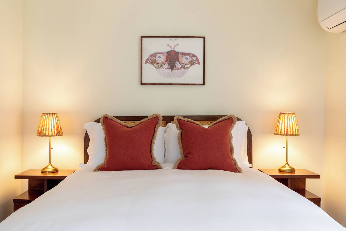 Un lit bien fait, avec du linge de lit blanc et deux oreillers rouges, flanqué de deux tables de chevet avec des lampes, dans une pièce où un tableau encadré orne le mur.