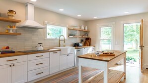 A modern kitchen with white cabinetry, wooden countertops, and a central island. There are windows providing natural light and a view of greenery outside. - Silas House (Lexington)