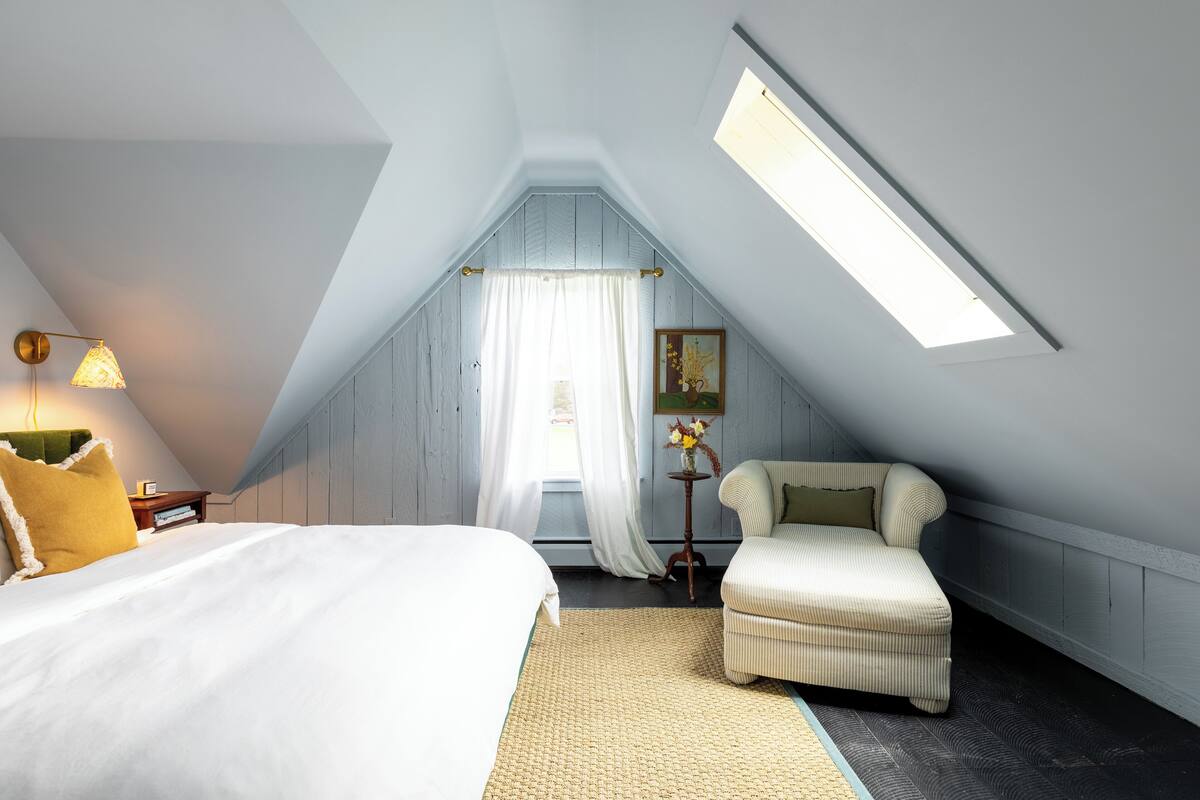 Une chambre avec un plafond en pente, un lit, une chaise, une petite table avec un vase et une fenêtre avec des rideaux.