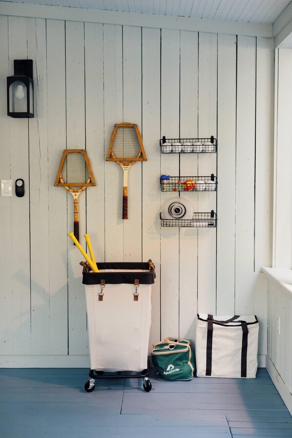 A room with a white wooden wall, a black wall-mounted light, and a rack holding tennis rackets and a bag. - Silas House (Lexington)