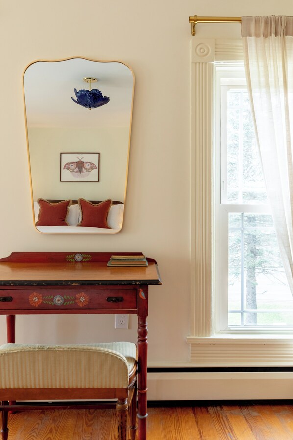 Una habitación con una mesa de madera, una silla, un espejo, un ventanal con cortinas y una obra de arte con una mariposa enmarcada.
