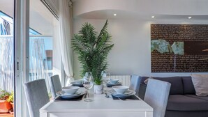 Een eetruimte met een witte tafel voor vier personen, omringd door grijze stoelen. Een grote potplant staat in het midden, en een grijze bank staat tegen de muur.