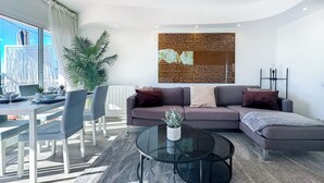 Een moderne woonkamer met een hoekbank, een glazen salontafel en een groot raam met gordijnen.